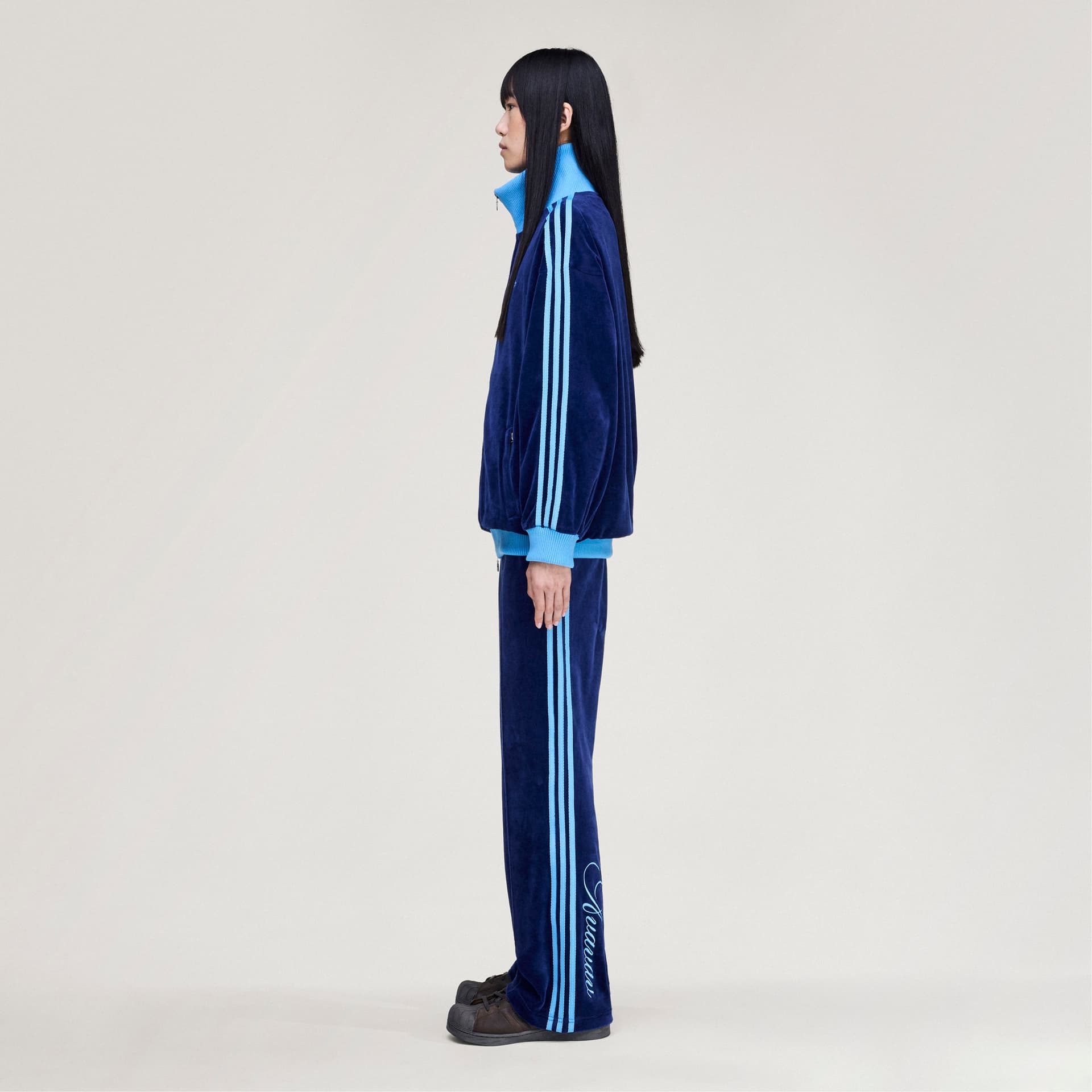 Side profile of a model wearing the adidas Originals by Avavav Velvet Track Jacket in dark blue with light blue stripes.