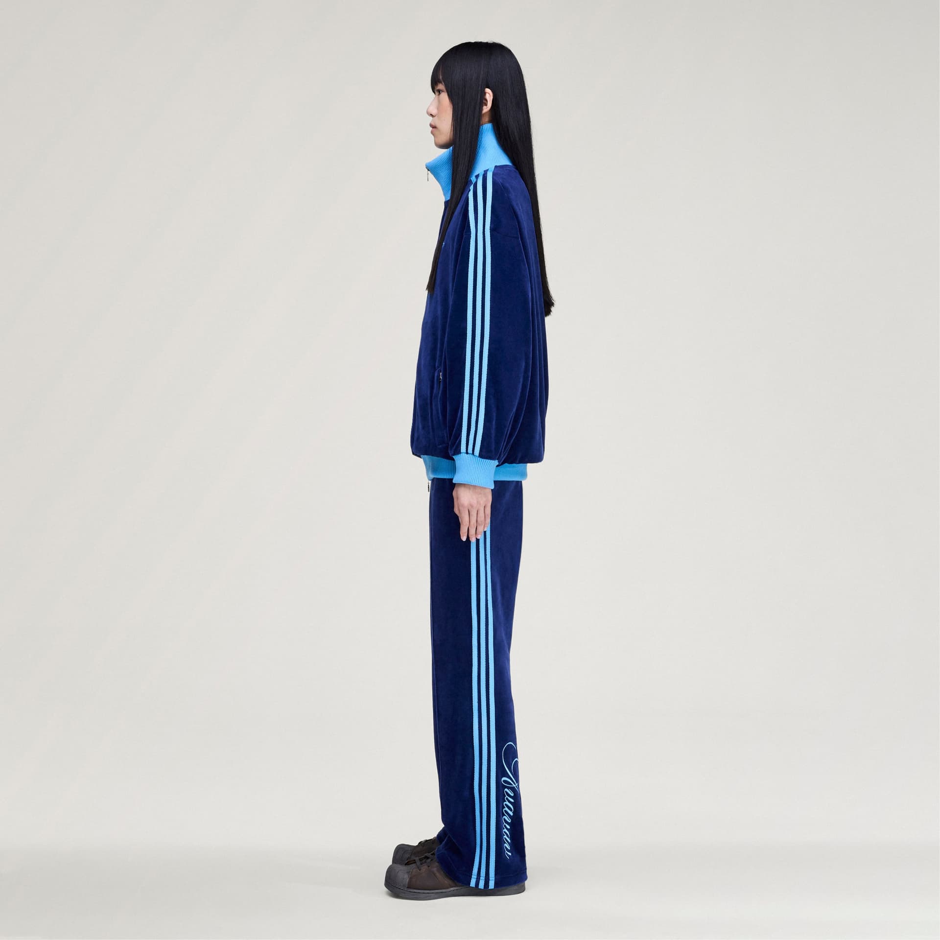 Side view of a model wearing the adidas Originals by Avavav Velvet Track Pant and jacket in navy with light blue stripes.