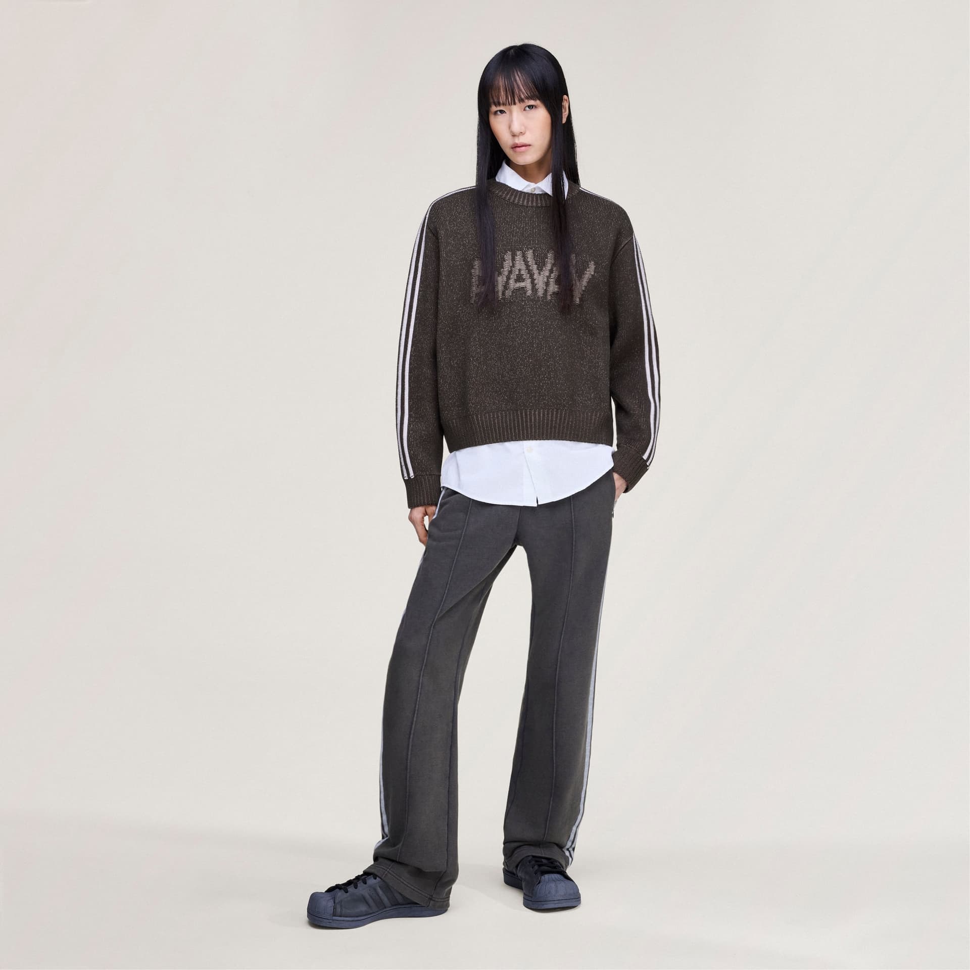 A model wearing the adidas Originals by Avavav Crewneck Sweater in dark brown, over a white shirt with matching trousers.