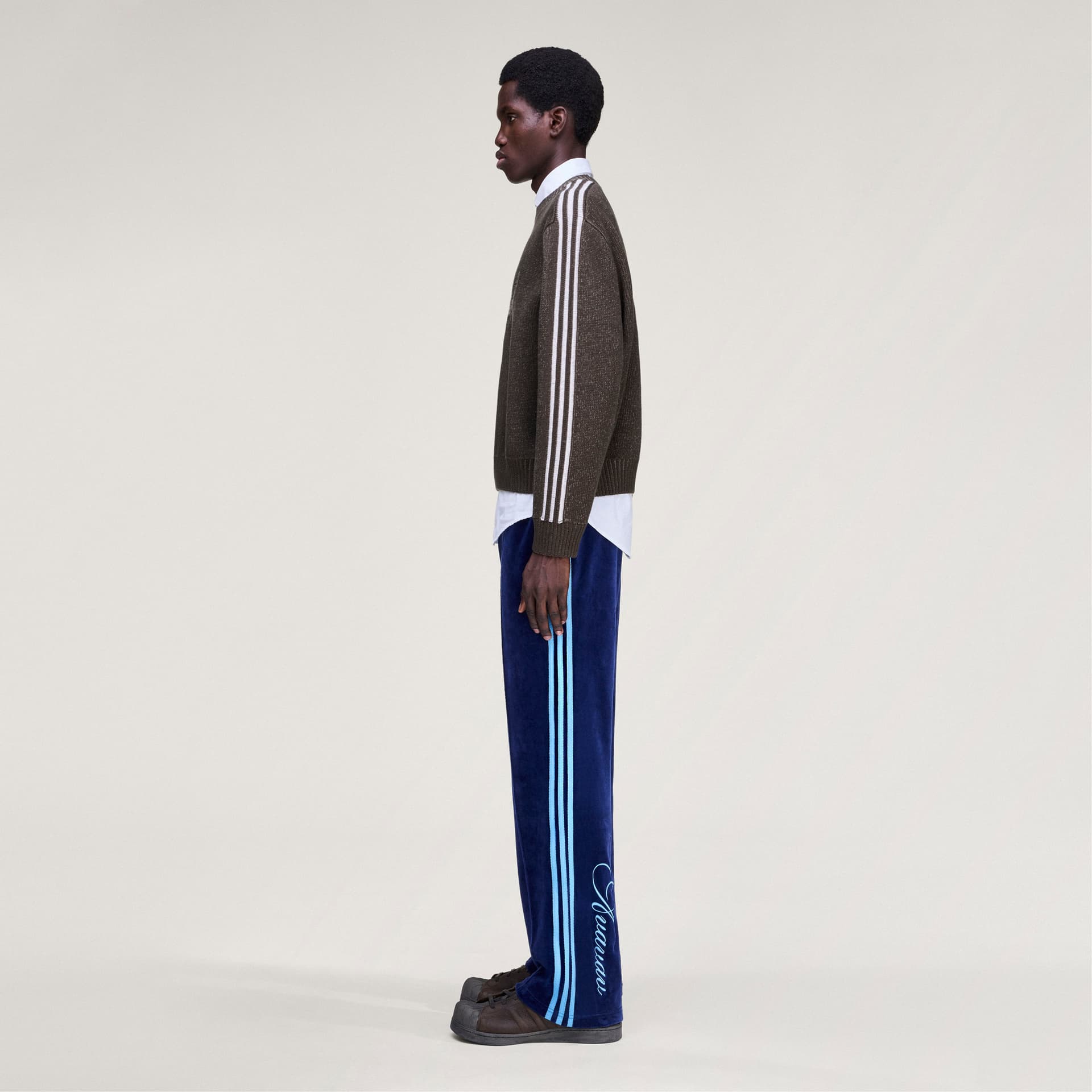 Side profile of a model wearing the adidas Originals by Avavav Crewneck Sweater in brown with white three-stripe detailing.