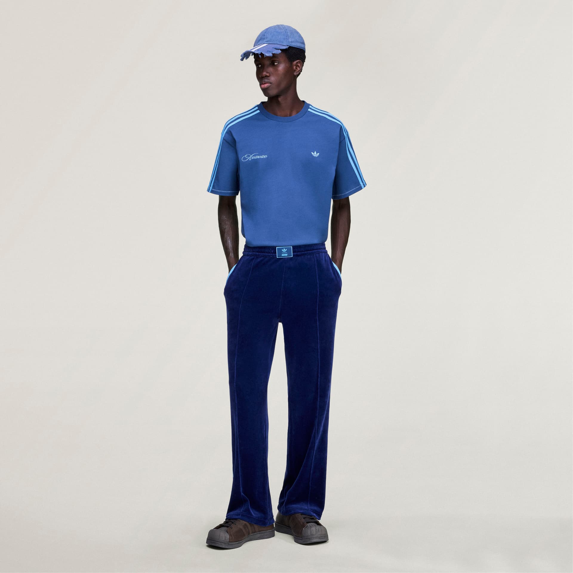 Full-length shot of a model wearing the blue adidas Originals by Avavav T-shirt, styled with dark blue velour pants.