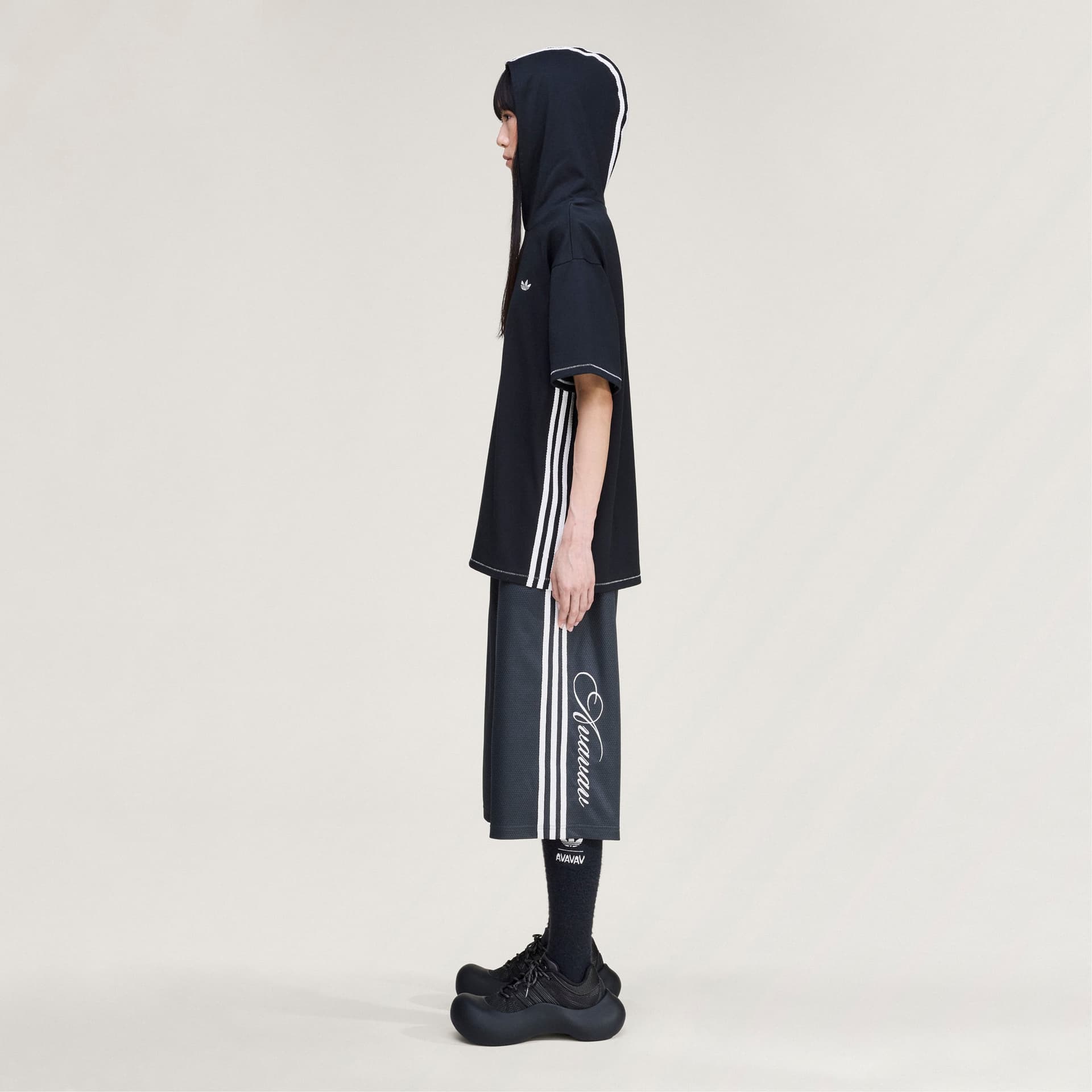 Side profile view of a model wearing the black adidas Originals by Avavav Hooded T-shirt with its signature white stripes.