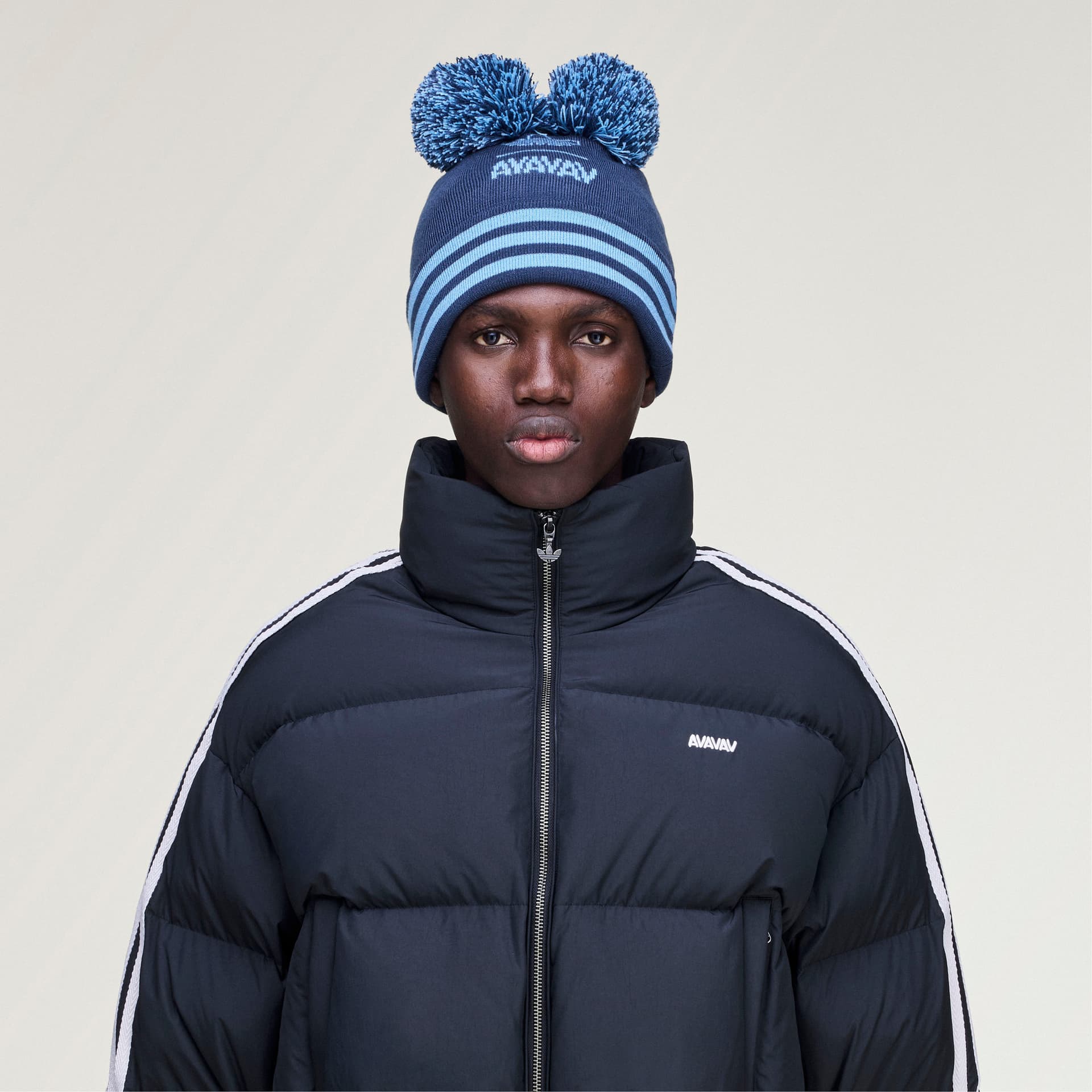 Front view of a model wearing the blue adidas Originals by Avavav beanie with two large pom-poms and light blue stripes.