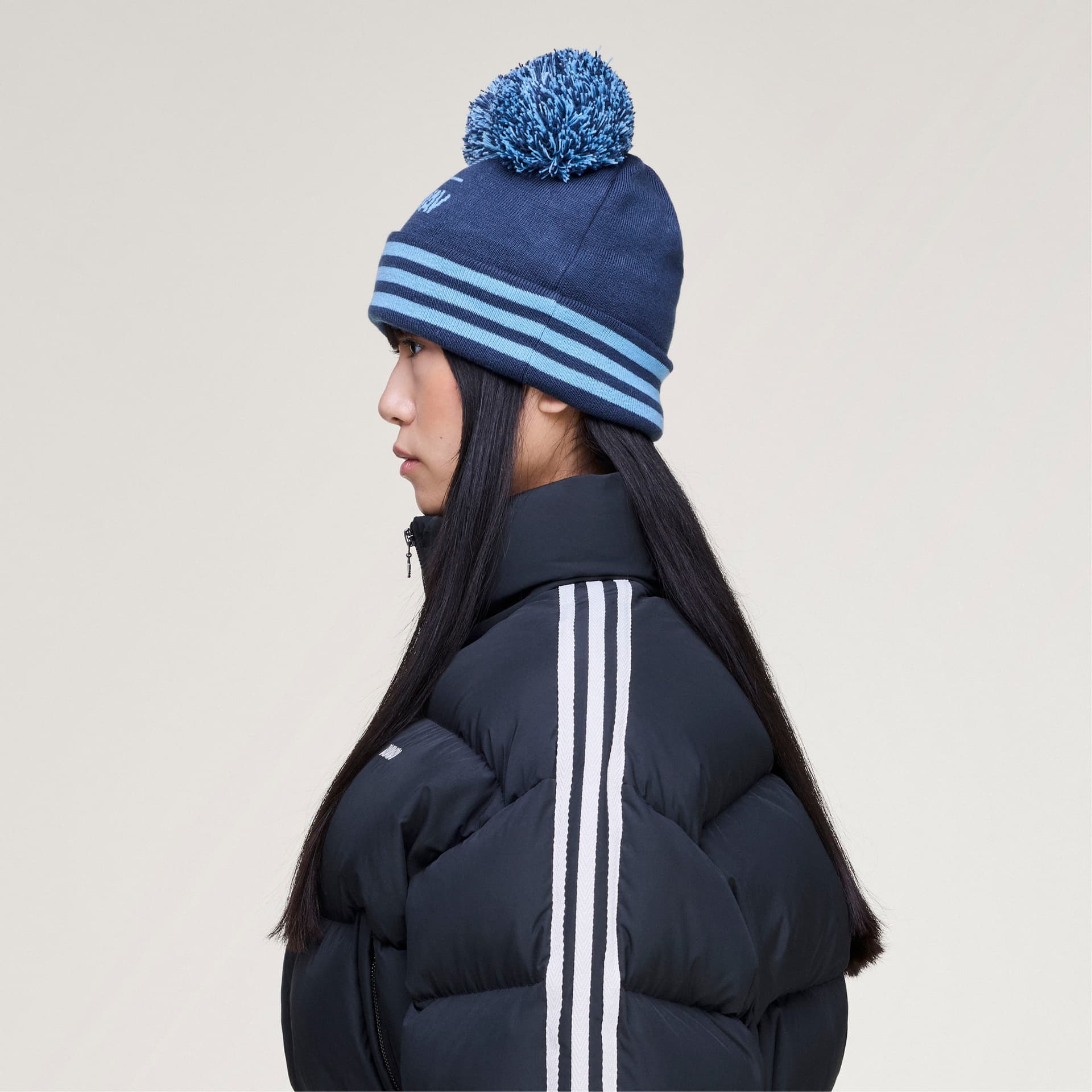 Side profile of a model wearing the blue adidas Originals by Avavav Pom Pom Beanie with a matching pom-pom and striped cuff.