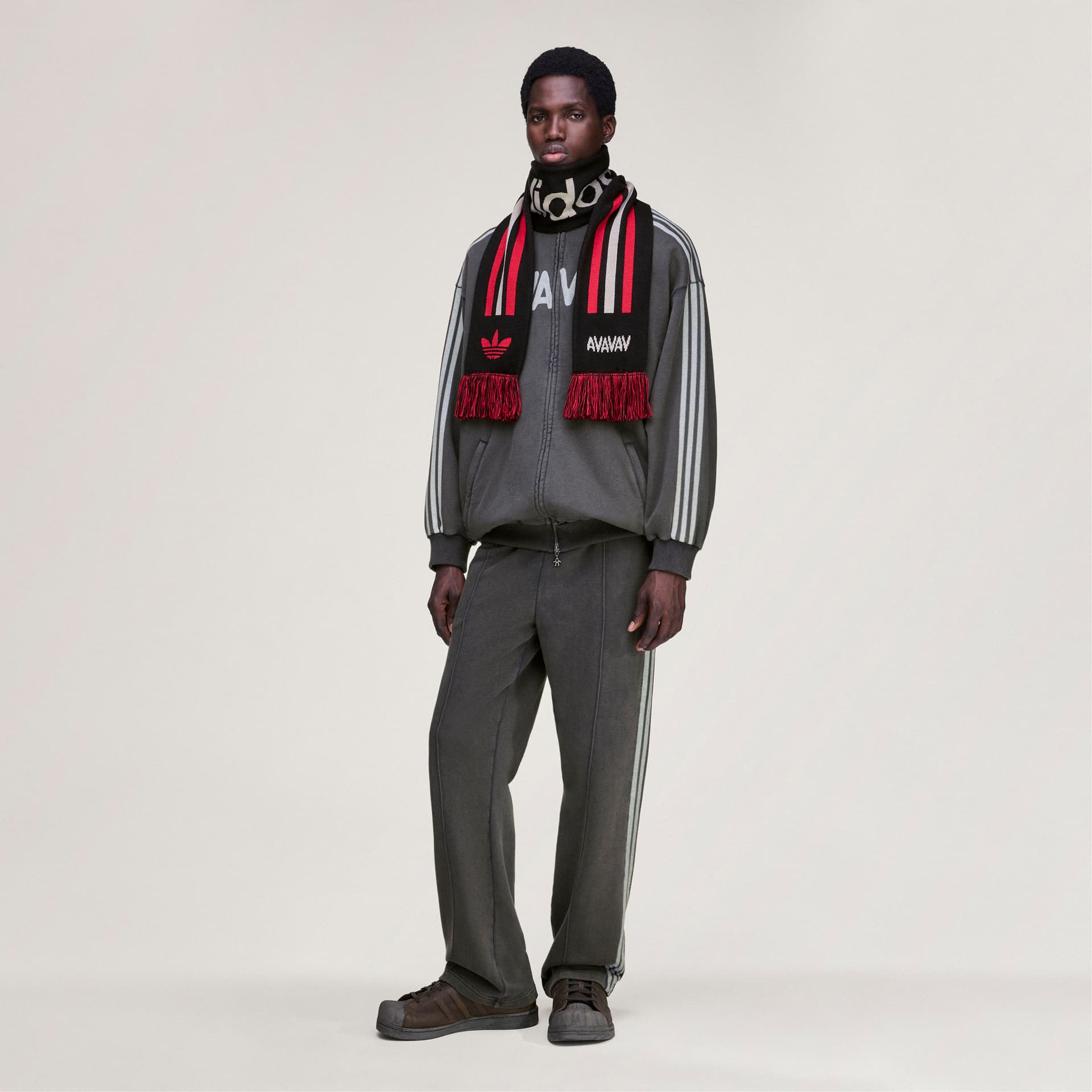 The adidas Originals by Avavav Scarf in black with red fringe, shown on a model wearing a coordinating grey tracksuit.